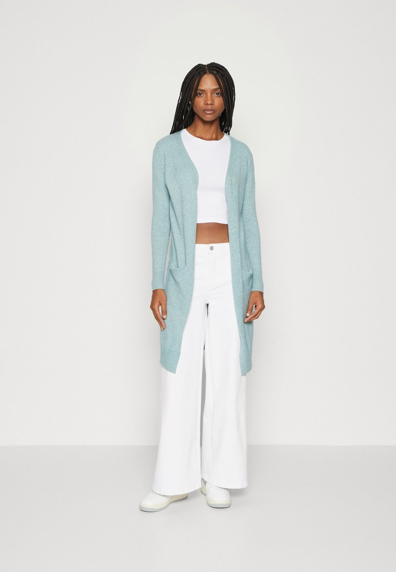 Lyseblå lang cardigan laget av mykt materiale, med åpen front, to sidelommer, og parret med en hvit crop top og vide bukser.