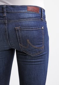 Jeans de denim azul oscuro con una forma ajustada, que presentan una etiqueta de cuero marrón de LTB en la cintura y costuras amarillas en contraste en los bolsillos.