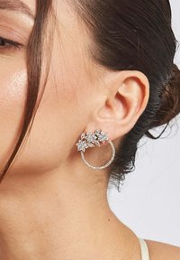 Guldörhängen i hoops design med blommönster prydda med klara strassstenar. Ytan är slät med en glänsande finish som förbättrar synligheten.