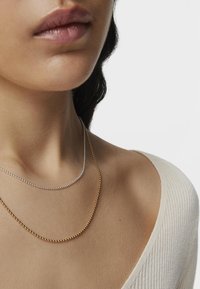 Dos collares en capas: una cadena de cuentas plateadas se encuentra sobre una cadena de cuentas doradas. El modelo lleva una camiseta de color claro con relieve.