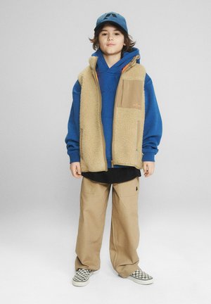 Dziecko ubrane w niebieską bluzę z kapturem, beżową kamizelkę polarową, beżowe spodnie, niebieską czapkę i kraciaste wsuwane buty, stojące na szarym tle.