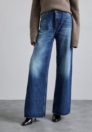 Person trägt hochtaillierte, weit geschnittene blaue Jeans, braunen Langarmpullover und schwarze Stiefel mit Absatz auf grauem Boden und weißer Wand.