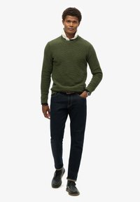 Grüner strukturierter Pullover, dunkle Jeans und schwarze Schuhe. Der Kragen eines neutralen Hemdes ist unter dem Pullover sichtbar. Das Model steht mit den Händen in den Taschen.