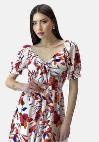 Vestido de estampado floral en tela blanca con patrones rojos, azules y naranjas. Presenta un escote cuadrado, mangas abullonadas y cintura fruncida.