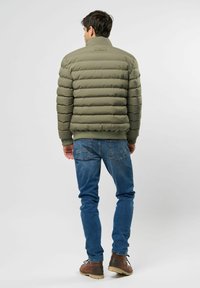Muškarac odjeven u maslinasto zelenu puffer jaknu, plave traperice i smeđe čizme stoji okrenut leđima prema jednostavnom sivoj pozadini.