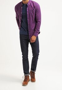 Rutig skjorta med knappar i lila och blått över en marinblå T-shirt; mörka jeans i denim; bruna snörskor med en texturerad yta.