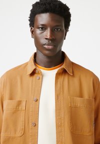 Braune Knopfjacke mit zwei Brusttaschen, getragen über einem cremefarbenen Hemd. Der Stoff hat eine strukturierte Optik.