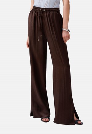 Donna che indossa pantaloni a gamba larga marrone scuro con coulisse in vita e piccole aperture laterali, abbinati a tacchi neri con punta aperta e braccialetto argentato.