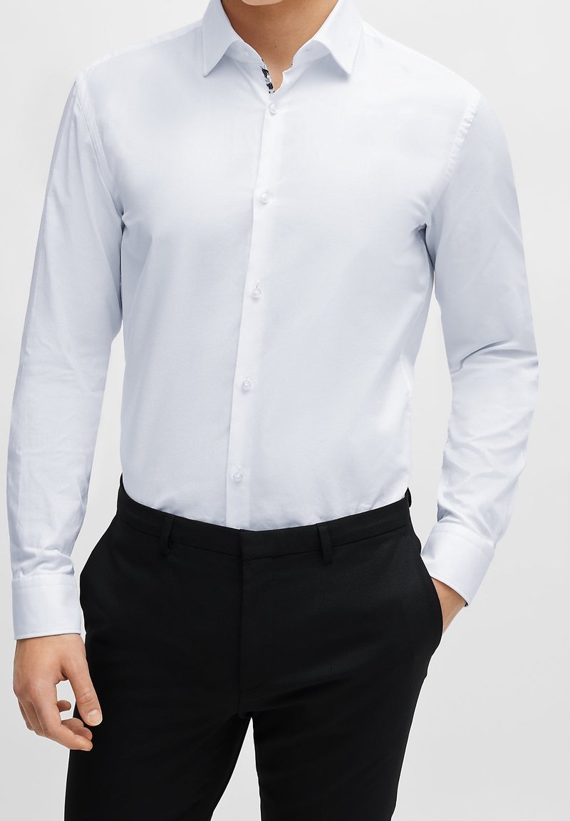Homme portant une chemise blanche ajustée à manches longues boutonnée, rentrée dans un pantalon noir, une main dans la poche.