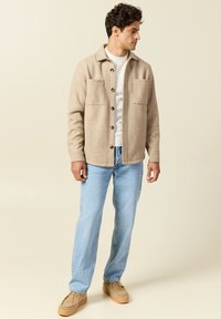 Giacca camicia in lana color beige chiaro con due tasche frontali, chiusura a bottone e colletto. Accompagnata da jeans blu chiaro e scarpe beige.