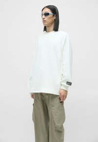 Hvid oversized sweatshirt med rund hals og ribbede manchetter, parret med lyse khaki cargo bukser med sidelommer. Modellen bærer grå solbriller.