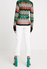 Kvinna som bär en långärmad topp med vågigt mönster i grönt, brunt, svart och vitt, vita upprullade byxor och gröna klacksandaler med blockklack.