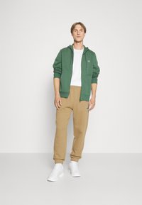 Grüne Zip-Hoodie über einem weißen T-Shirt, kombiniert mit beigen Hosen. Mit elastischen Bündchen und Taillenbund, dazu weiße Sneaker.