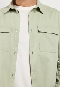 Camisa de botones verde claro con bolsillos de solapa, llevada abierta sobre una camiseta blanca de cuello redondo en la parte superior del torso de una persona.