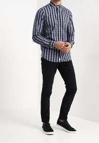 Uomo che indossa una camicia a quadri blu e nera, pantaloni neri slim-fit e sneaker nere con suola bianca, in piedi contro uno sfondo bianco.