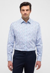 Camicia bianca con un motivo a foglie blu e verdi, maniche lunghe e colletto classico, abbinata a pantaloni blu navy.