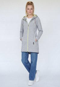 Žena s blond vlasmi v copu, oblečená v svetlosivej kapucňovej bunde na zips, modrých džínsach a bielych teniskách, stojaca s rukami v nohaviciach.