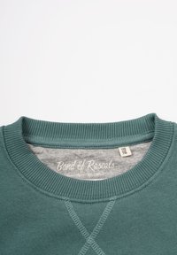Grøn sweatshirts med ribbet rund hals og lys grå inderside. Har en krydsstingdetalje foran. Brandmærket er synligt.