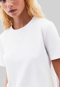 T-shirt en coton blanc à manches courtes, avec un col rond et des détails de couture subtils le long de l'encolure. Texture lisse et unie.