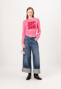 Suéter corto rosa con el texto "JOOP! JEANS", combinado con unos vaqueros de pierna ancha en azul con puños de mezclilla en contraste y botas negras de tacón.