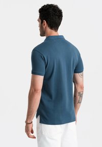 Pánská tmavě modrá polo tričko s krátkými rukávy, strukturovaná látka, klasický límec a postranní rozparky, nošeno s bílými kalhotami.