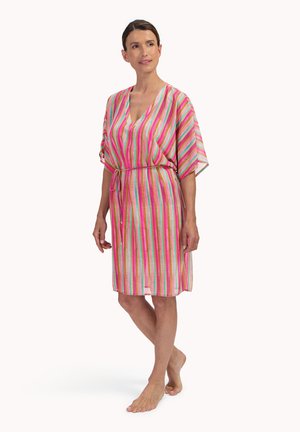 Vrouw die een knie-lange, kleurrijk gestreepte jurk draagt met een V-hals en losse mouwen, staand op blote voeten in een ontspannen houding met een strikceintuur in de taille.