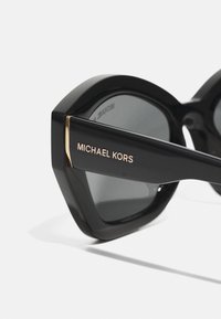 Melnas taisnstūra formas saulesbrilles ar pelēkiem stikliem, izceļoties ar izteiksmīgu stūrainu dizainu, spīdīgu apdari un zelta akcentu ar "MICHAEL KORS" zīmolu.