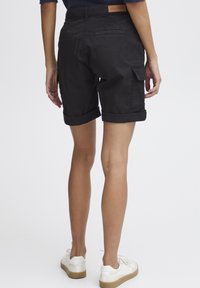 Person som bär svarta uppvikta cargoshorts, vita sneakers och en marinblå stickad topp, visad bakifrån mot en vit bakgrund.