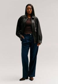Jeune femme debout les mains dans les poches, portant une veste en cuir noire, un haut marron foncé, un pantalon large bleu foncé et des chaussures marron à talons.