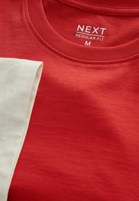 T-shirt rouge col rond avec une étiquette blanche « NEXT Regular Fit M » imprimée à l'intérieur du col, partiellement recouverte par un tissu beige plié.