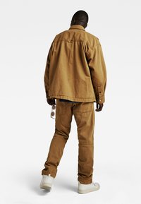 Bruine katoenen oversized jas met een kraag, gecombineerd met bijpassende baggy broek en witte high-top sneakers. Outfitaccessoires: lichtgekleurde zonnebril.