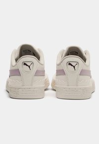 Bílé tenisky s texturovanou podrážkou, zdobené lilákovým proužkem a logem Puma na patě. Design zahrnuje zvlněné akcenty na zadní straně.