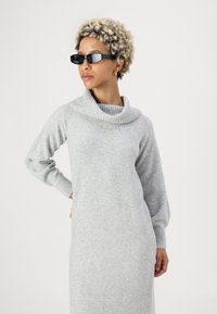 Vestido de malha cinza com decote drapeado e mangas longas. Textura suave, punhos canelados e design ajustado. A modelo usa óculos de sol retangulares pretos.