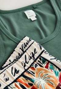 Grünes geripptes Tanktop kombiniert mit einem plissierten Rock, der abstrakte bunte und schwarze Schriftmuster auf cremefarbenem Hintergrund zeigt.