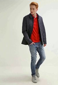 Tânăr cu părul roșu îmbrăcat cu o jachetă neagră matlasată, un tricou polo roșu, blugi de culoare albastru deschis și adidași gri, stând în fața unui fundal deschis uniform.