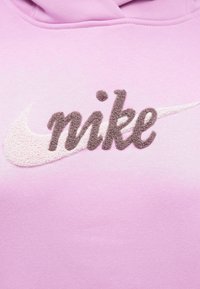 Rosa hettegenser med en stor, strukturert hvit Nike swoosh-logo og brodert mørkegrå "nike"-tekst i et mykt, plysjete stoff.