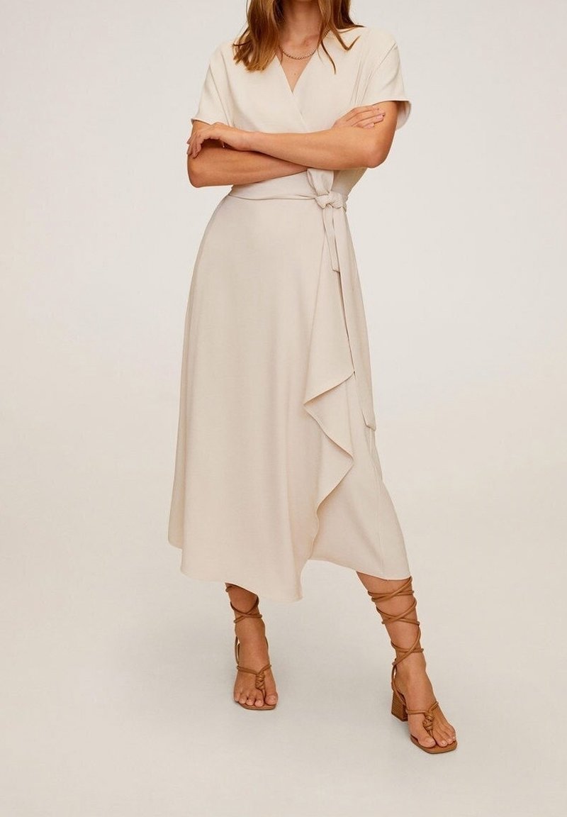 Robe croisée beige clair à manches courtes, en tissu doux, avec un ourlet asymétrique et un détail de ceinture nouée. Associée à des sandales échancrées couleur tan.