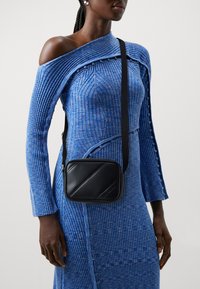 Borsa a tracolla in finta pelle nera con un design diagonale, caratterizzata da un profilo sottile e una tracolla rimovibile, indossata con un vestito blu a coste.