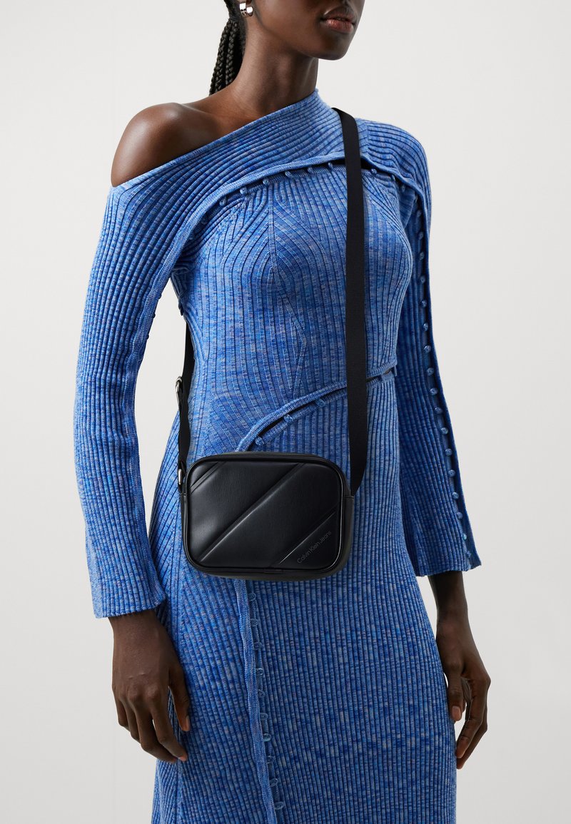 Borsa a tracolla in finta pelle nera con un design diagonale, caratterizzata da un profilo sottile e una tracolla rimovibile, indossata con un vestito blu a coste.
