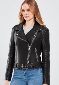 Zwart leren motorjack met zilveren studs langs de kraag en ritsen, voorzien van een ceintuur in de taille en een aansluitend ontwerp.