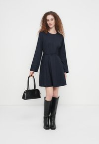 Φόρεμα σε σκούρο μπλε με λεπτές ρίγες, μακριά μανίκια, ζώνη στη μέση, φαρδιά φούστα· συνδυασμένο με μαύρα μποτάκια μέχρι το γόνατο και μικρή μαύρη τσάντα.