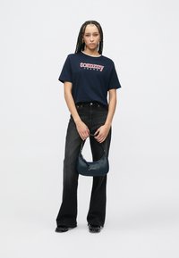 Tengerészkék póló "tommy" logóval, magas derekú fekete farmerrel párosítva. Egy sötétkék táskát tart a kezében, amelyen ezüst színű logó látható.