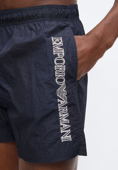 Μπλε σκούρο σορτς με ελαστική μέση, χέρι στην τσέπη, λευκό λογότυπο "Emporio Armani" κάθετα κεντημένο στο αριστερό μηρό.