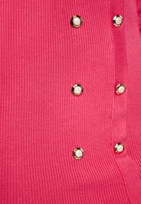 Top in tessuto rosa a coste con accenti esagonali dorati e centri bianchi, chiusure laterali con bottoni e texture liscia.
