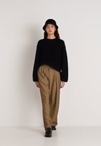 Μαύρο κοντό πουλόβερ, καφέ πλισέ παντελόνι και μαύρα παπούτσια. Το μοντέλο φοράει μαύρο καπέλο τύπου κάλπη, στέκεται μπροστά σε ανοιχτό φόντο.