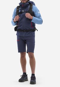 Marineblau und hellblaue Wanderjacke mit roten Akzenten, kombiniert mit marineblauen Shorts. Schwarzer Rucksack, Shorts mit einem Reißverschlussfach und dunklem Schuhwerk.
