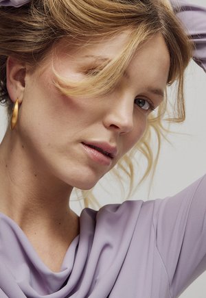 Donna bionda con capelli mossi indossa un orecchino a cerchio dorato e una maglia lavanda, con il braccio dietro la testa, guarda leggermente verso il basso di lato.