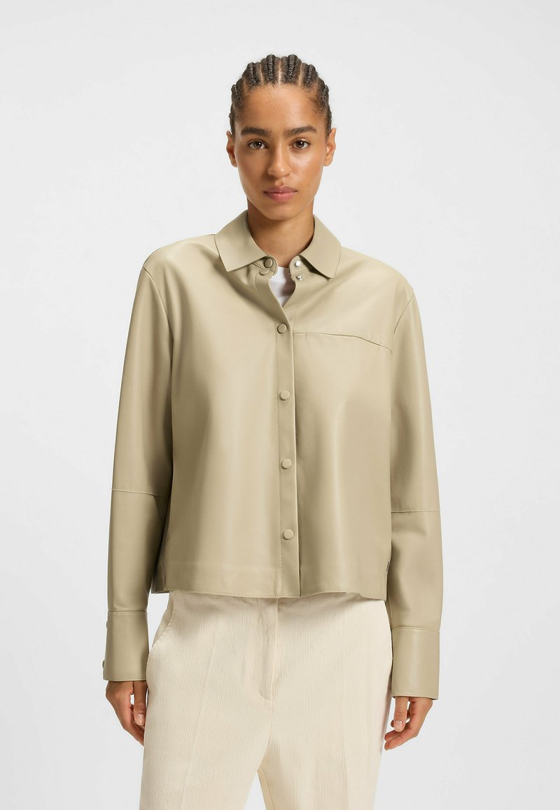Frau mit geflochtenem Haar, die eine beige, knöpfbare Lederjacke und hellfarbige Cordhosen trägt, vor einem einfarbigen Hintergrund.