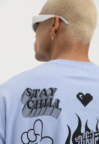 Person med kort blondt hår som har på seg hvite solbriller og en lyseblå skjorte med svart tekst "Stay Chill" og grafiske design på baksiden.