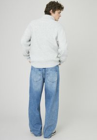 Pull en tricot gris avec un col montant, associé à un jean bleu à jambes larges avec poches arrière. Le modèle pose de dos sur un fond clair.